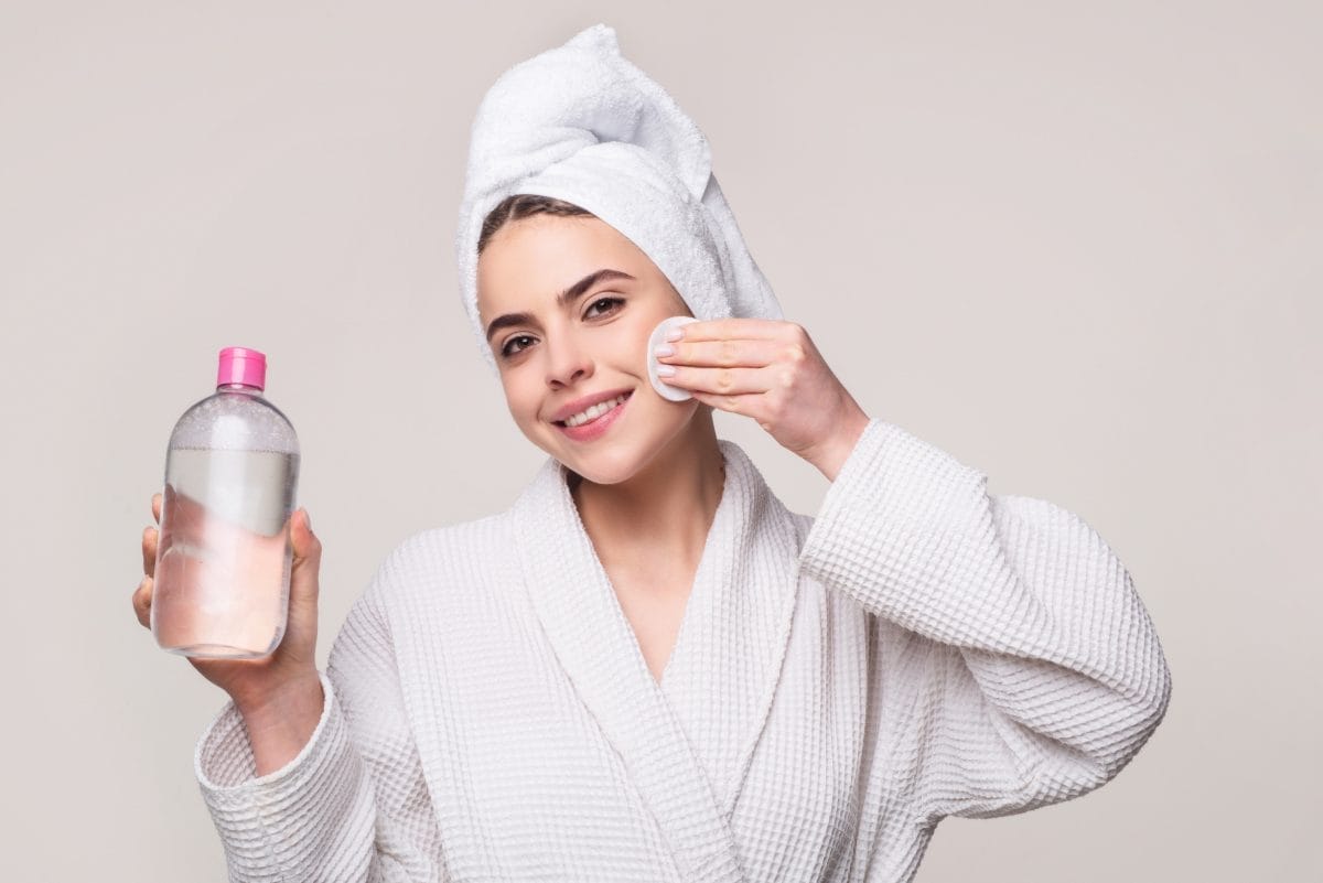 happy young woman in bathrobe with towel on head cleansing face with micellar water and cotton pad isolated on pink. girl with micellar water.