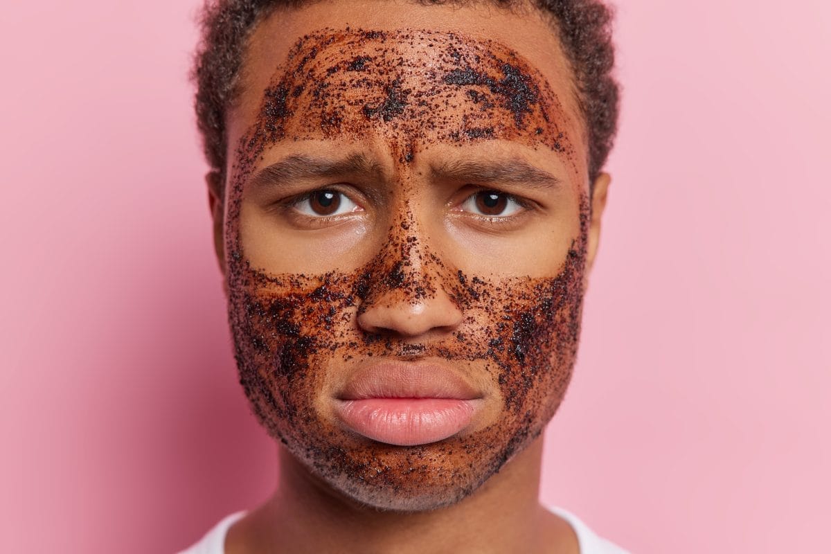 close up portrait of serious displeased man models with coffee scrub mask on his face frowns and has discontent expression takes care of skin condition isolated over pink background. beauty concept