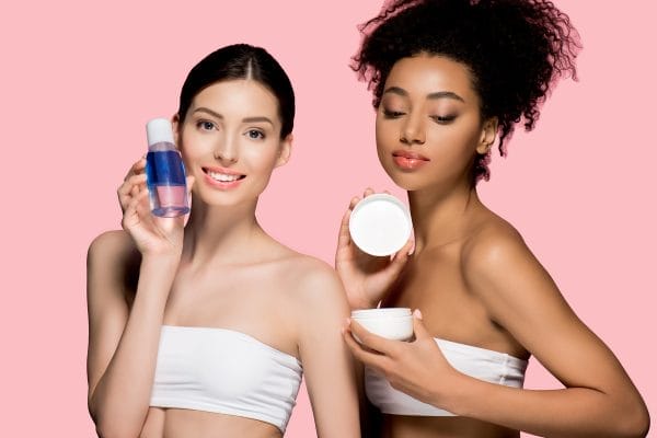 happy multiracial women holding cosmetic cream and makeup remover, isolated on pink