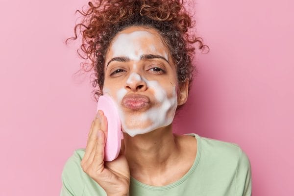 portrait of afro american woman with curly hair keeps lips folded washes face with sponge applies foam wants to have clean skin dressed in green jumper isolated over pink background. hygiene concept