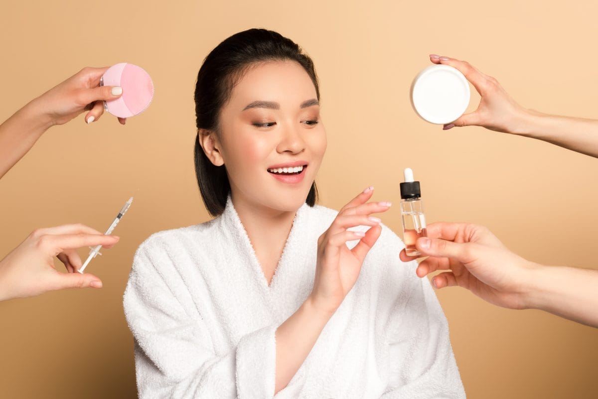 smiling beautiful asian woman in bathrobe near hands with facial cleansing brush, syringe, cosmetic