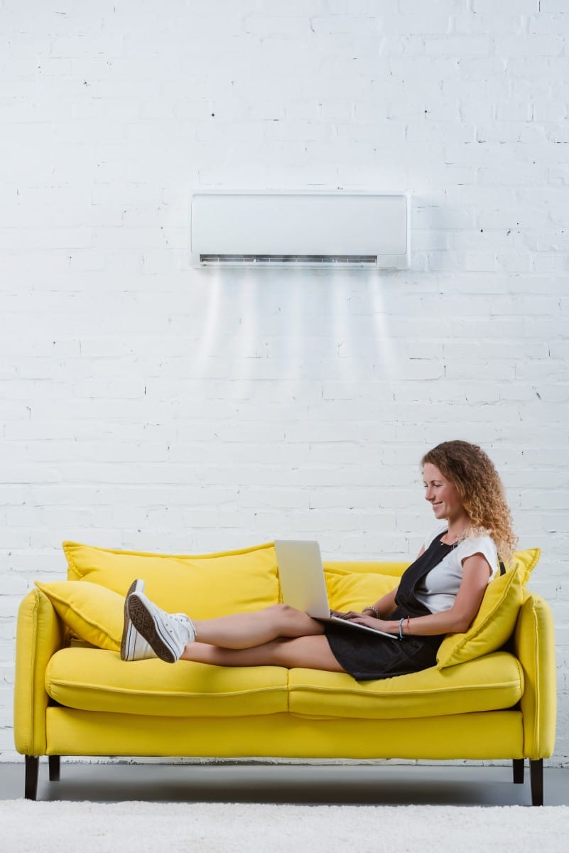 beautiful young woman working with laptop on couch under air conditioner hanging on wall