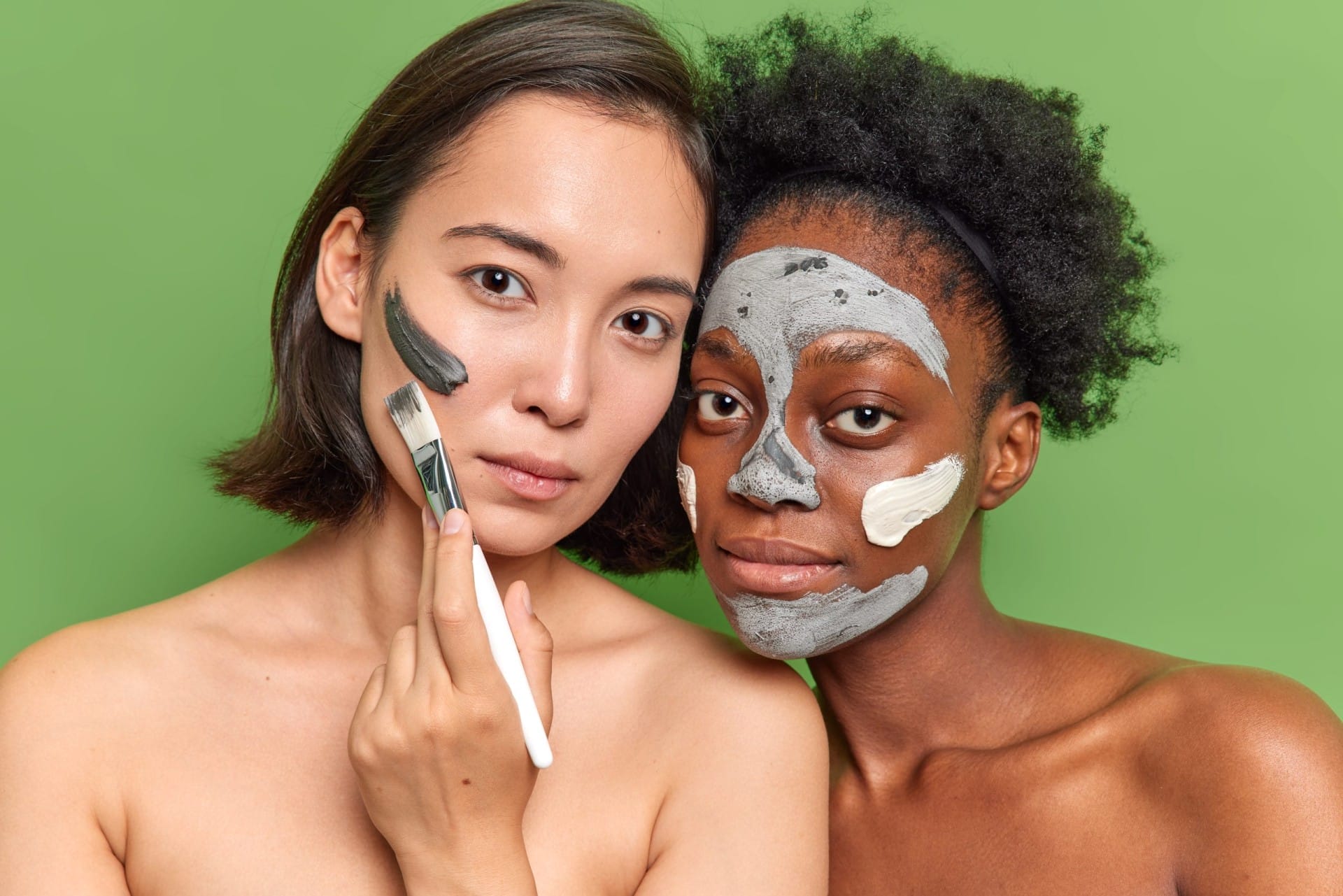 portrait of diverse young women look seriously at camera apply clay mask use cosmetic brush stand topless against vivid green background have delicate skin enjoy spare time for beauty treatments