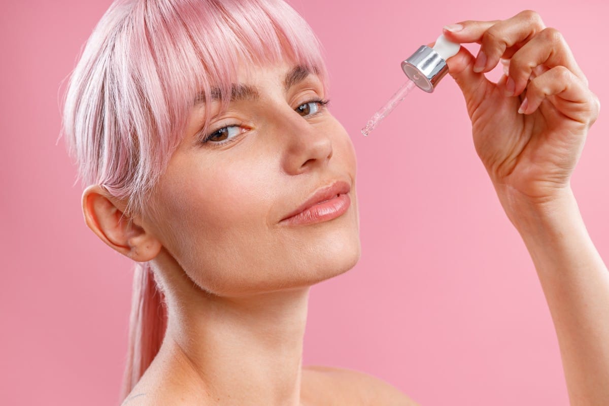 close up portrait of young woman with pink hair applying serum or hyaluronic acid on her facial skin using dropper isolated over pink background