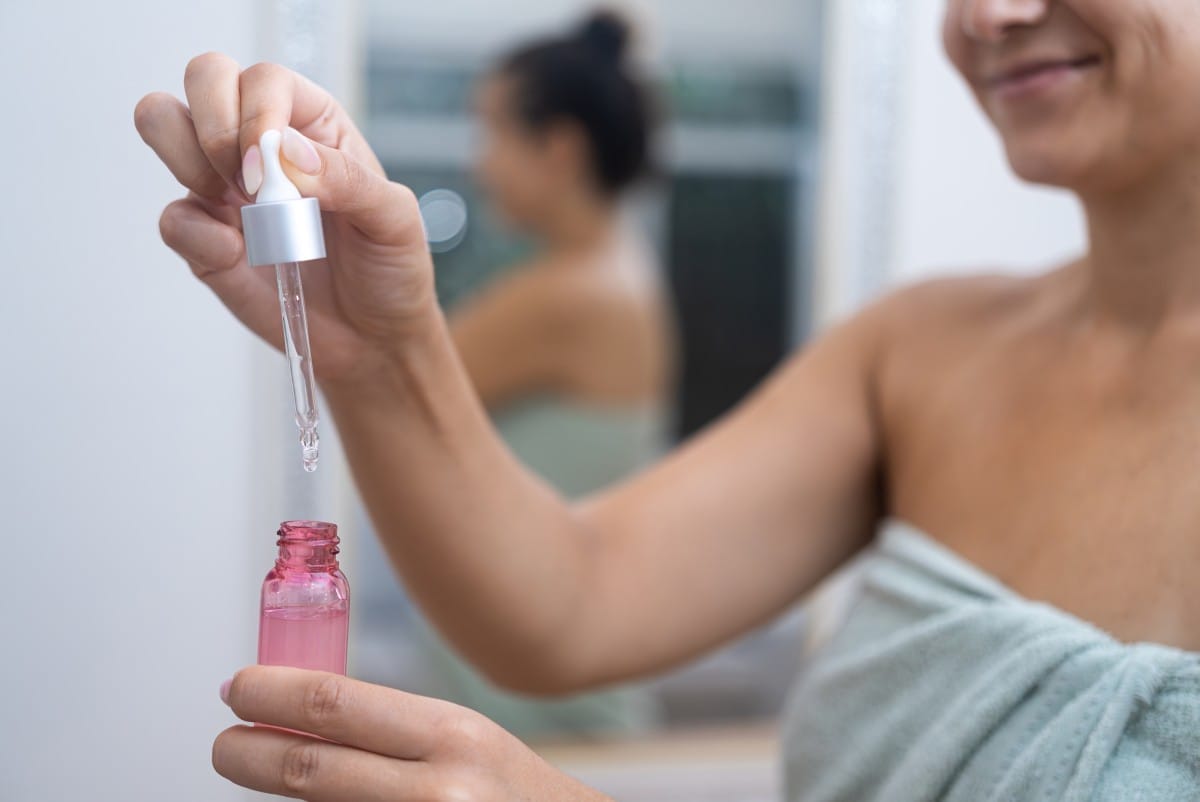 smiling woman with towel dripping serum over small bottle in bathroom lady showing hyaluronic acid