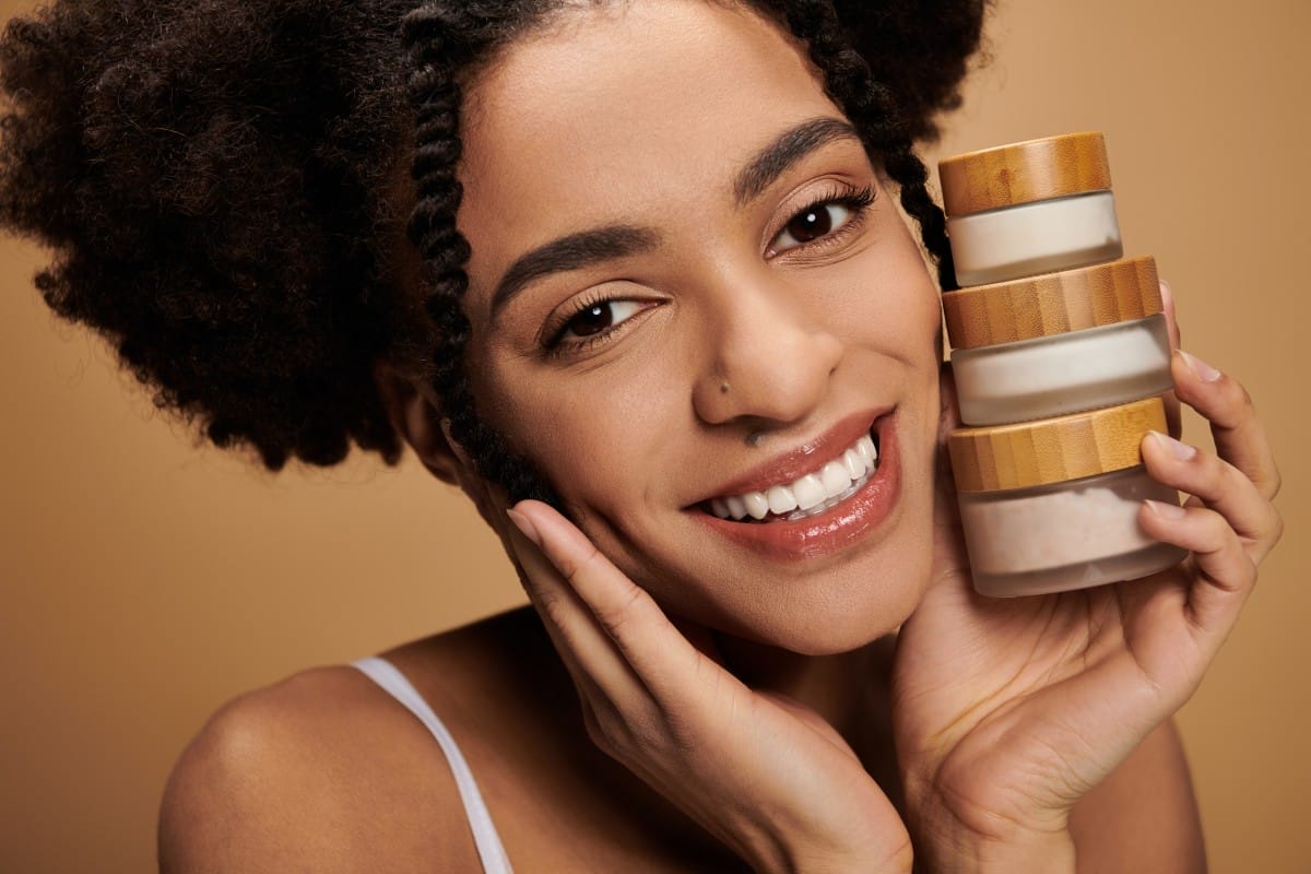 a young african american woman with a radiant smile holds skincare products against her face.