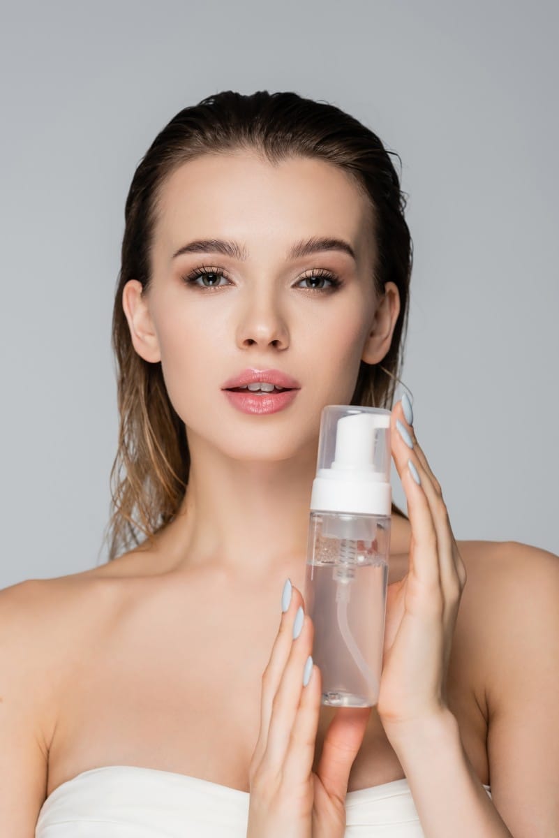 young woman with clean skin and wet hair holding face foam isolated on grey