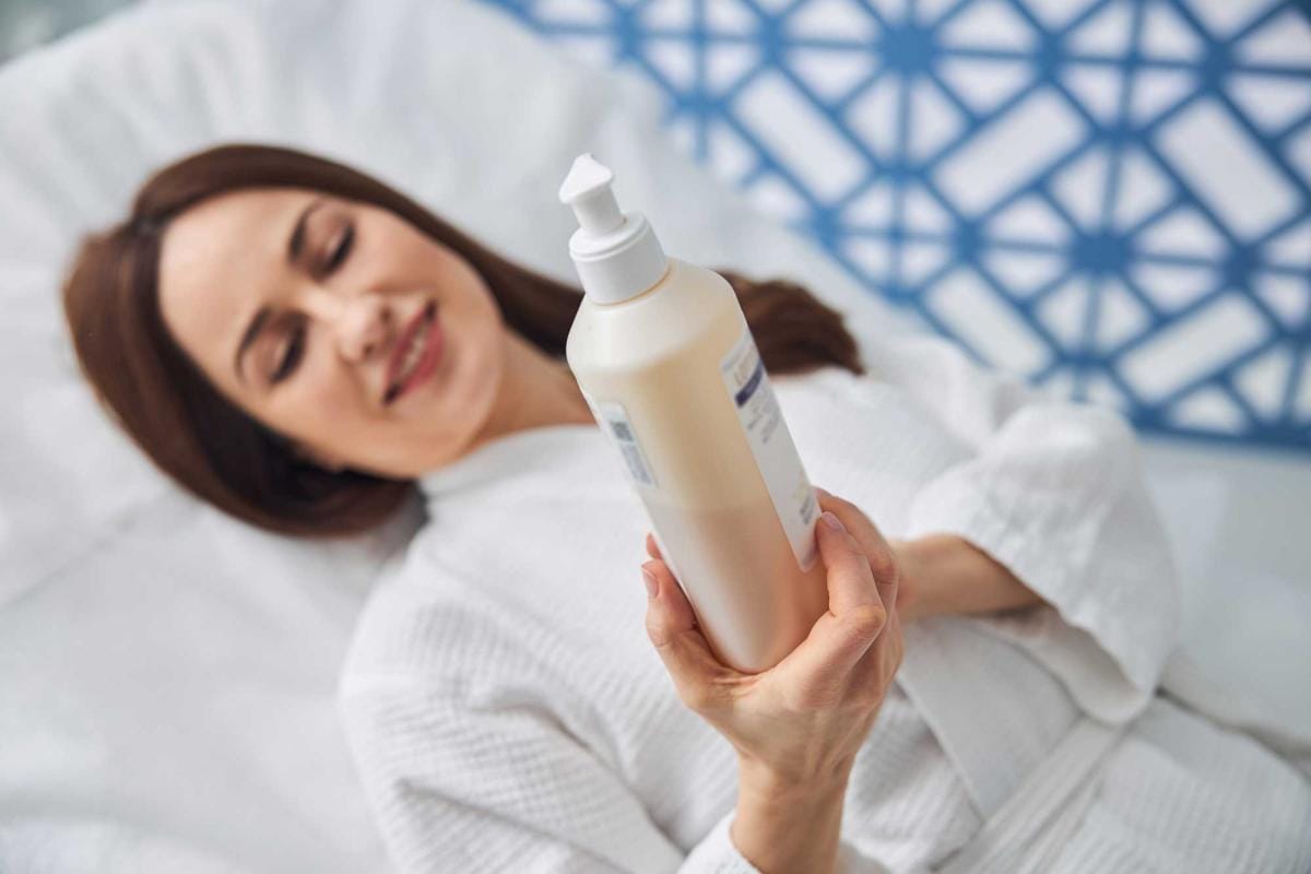 Woman holding skincare product while relaxing on bed