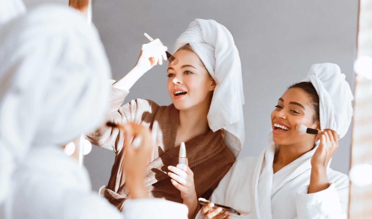 Women applying makeup and smiling in bathroom mirror