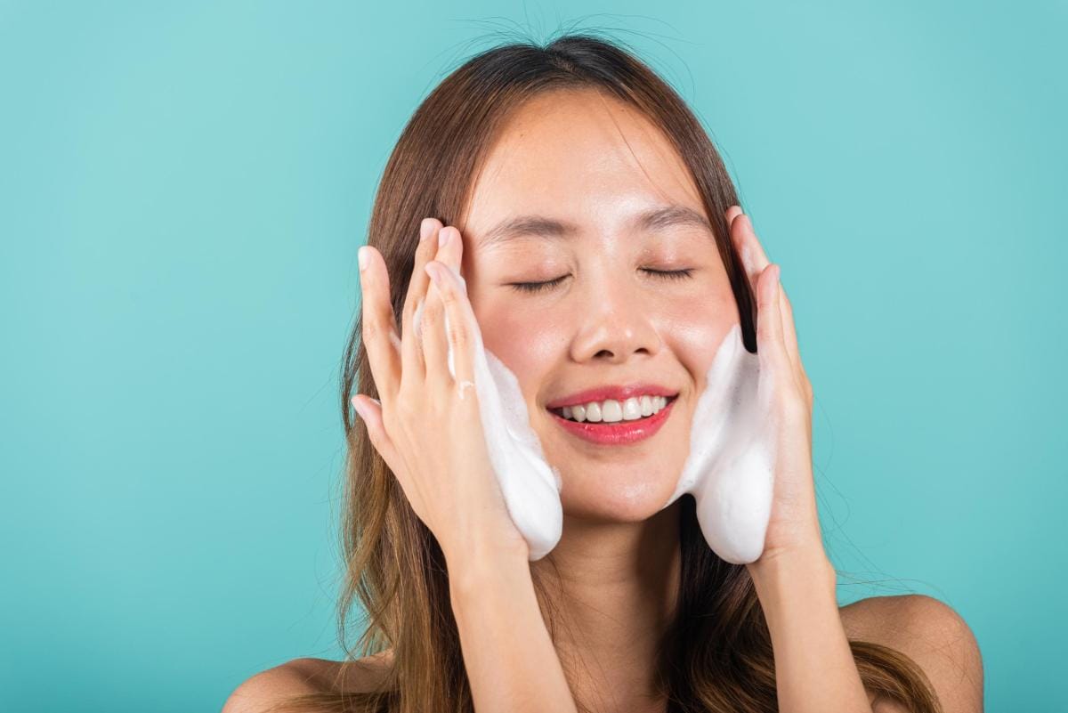 Woman happily applying facial foam, teal background.