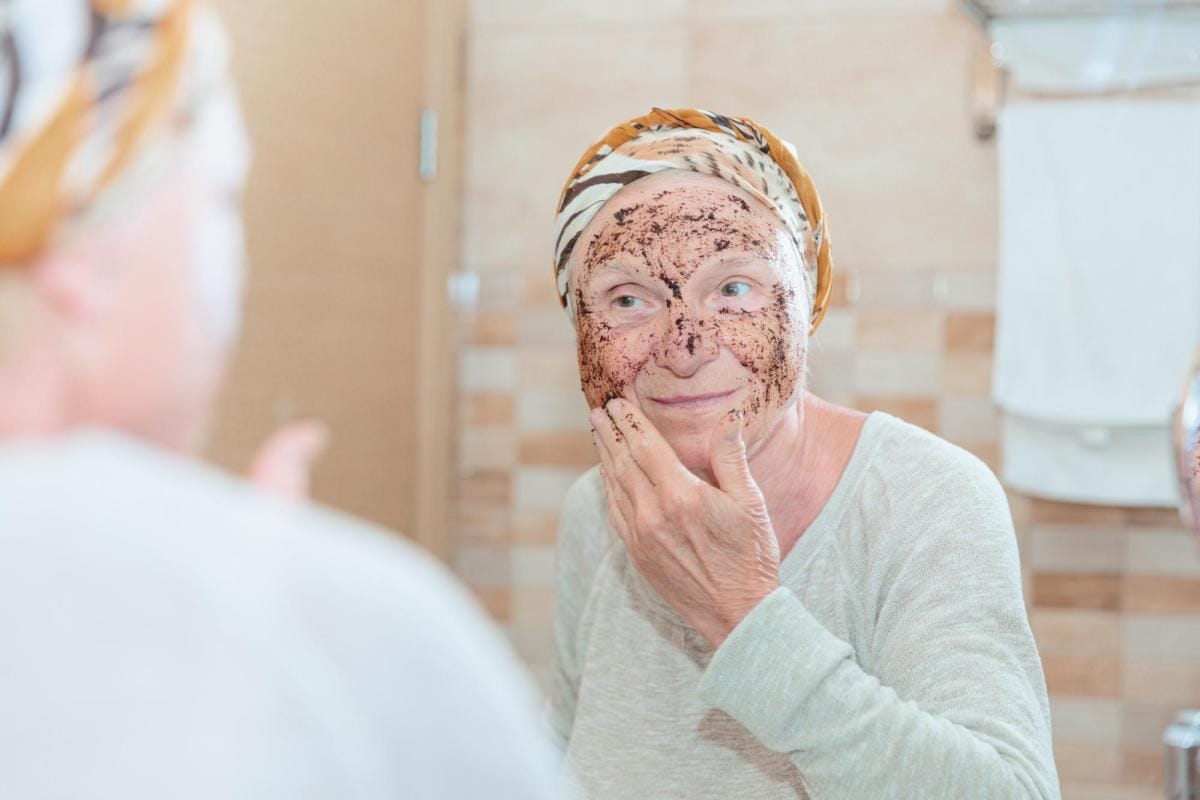 Elderly woman applying coffee scrub mask in bathroom mirror