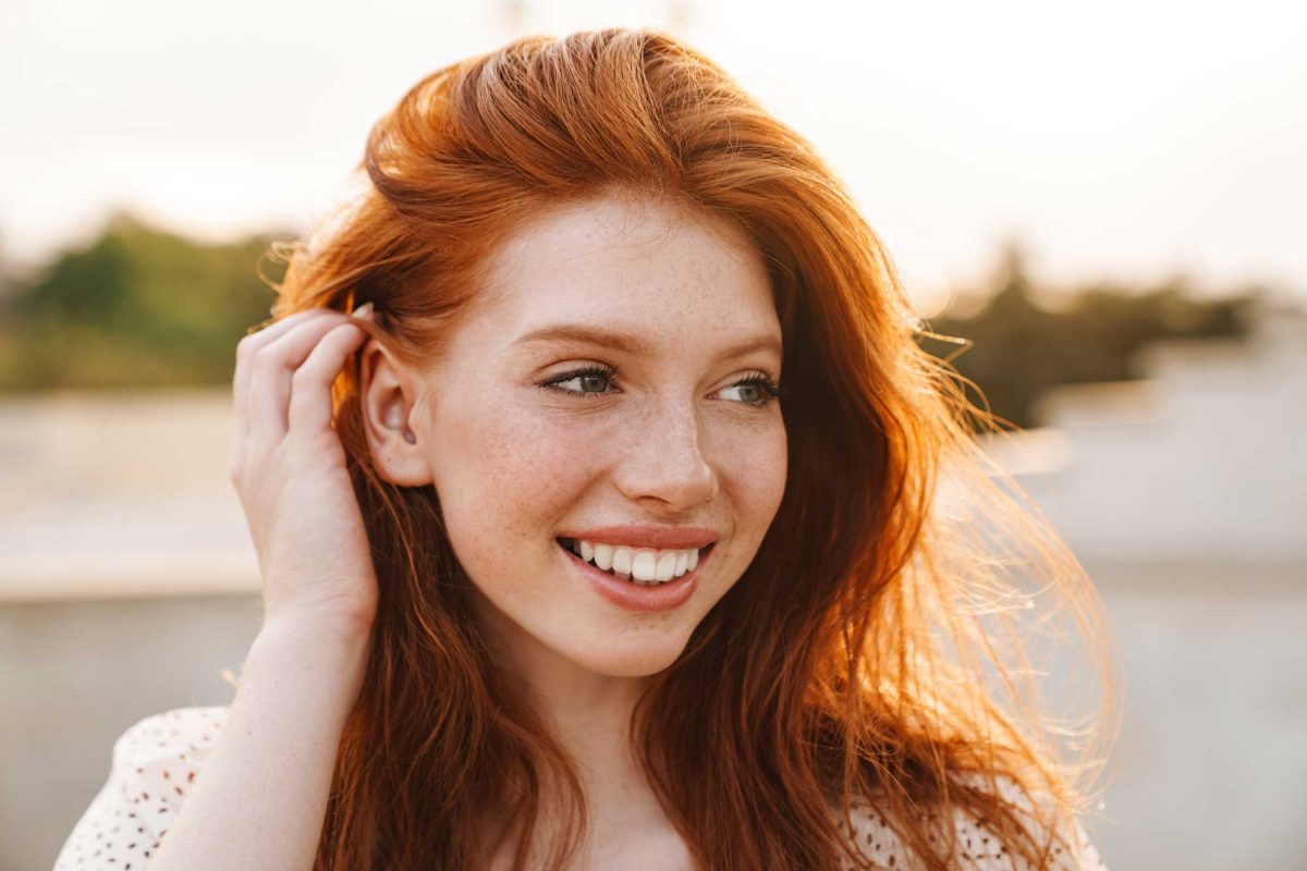 Smiling redhead woman touching hair in sunlight